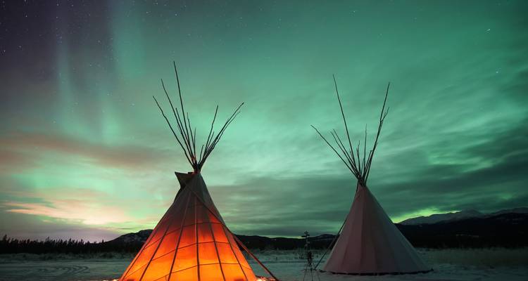 Twee gloeiende tipi's op besneeuwde grond onder een levendige groene aurora in de Arctische nacht.
