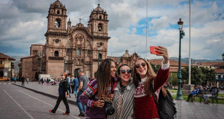 Tres mujeres con suéteres coloridos riendo y tomándose una selfie frente a la Catedral del Cusco en la plaza principal
