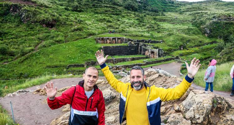 Dos viajeros felices con los brazos alzados frente a exuberantes ruinas incas en una ladera verde