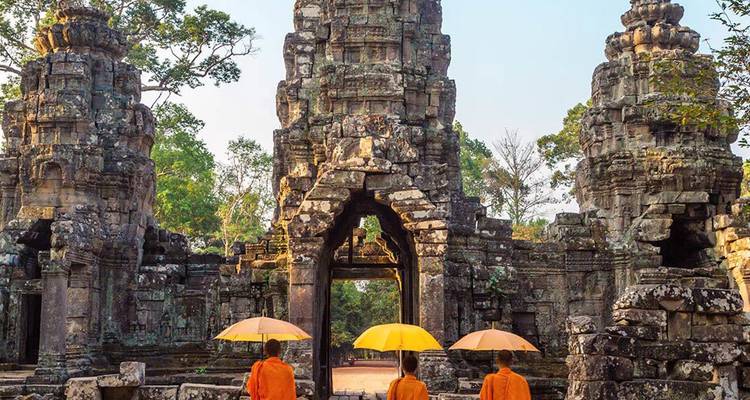 Drei orangegewandete Mönche mit gelben Regenschirmen gehen durch antike Khmer-Steintor-Ruinen.