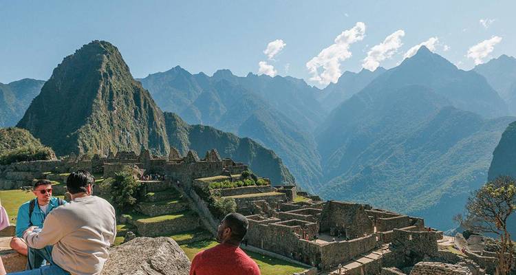 Panoramic view of Machu Picchu terraces framed by towering Andes peaks as travellers sit and admire the scene.