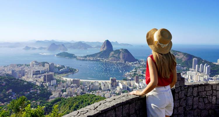 Young female traveler in straw hat gazes over Rio de Janeiro bay and Sugarloaf from a stone viewpoint.