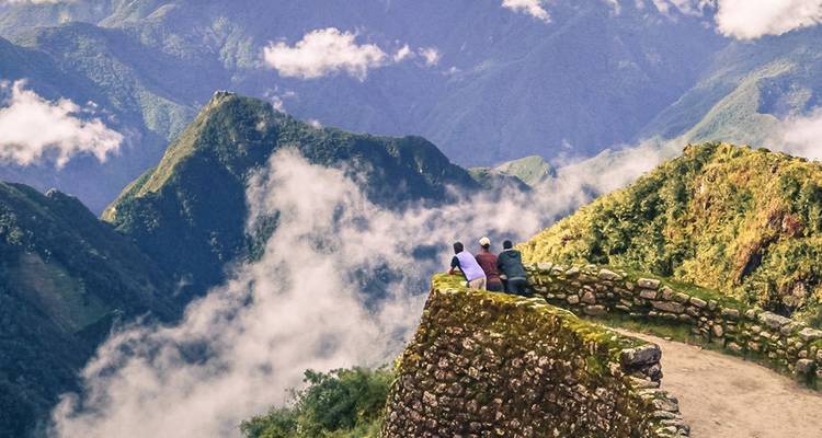 Three hikers rest on a mossy stone lookout high in the cloud-kissed Andes, Inca Trail below.