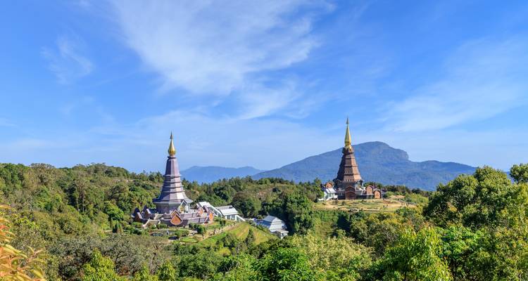 Twin pagodas with golden spires rising above lush gardens and forested mountains under a bright blue sky.