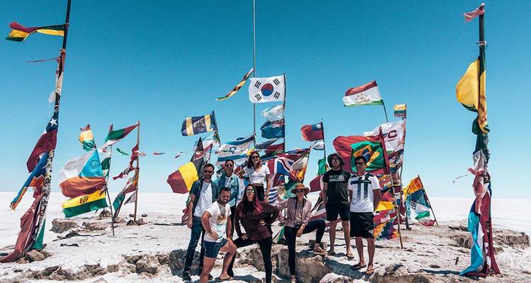 Een vrolijke groep reizigers die poseren tussen een cluster van kleurrijke nationale vlaggen op de uitgestrekte witte Uyuni zoutvlaktes onder een heldere blauwe hemel.