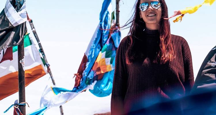 Mujer de pie en el salar de Uyuni junto a banderas internacionales coloridas bajo un cielo azul brillante