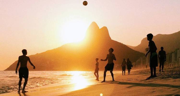 Las siluetas juegan fútbol playa al atardecer en la Playa de Ipanema con los picos de Dois Irmãos de fondo.