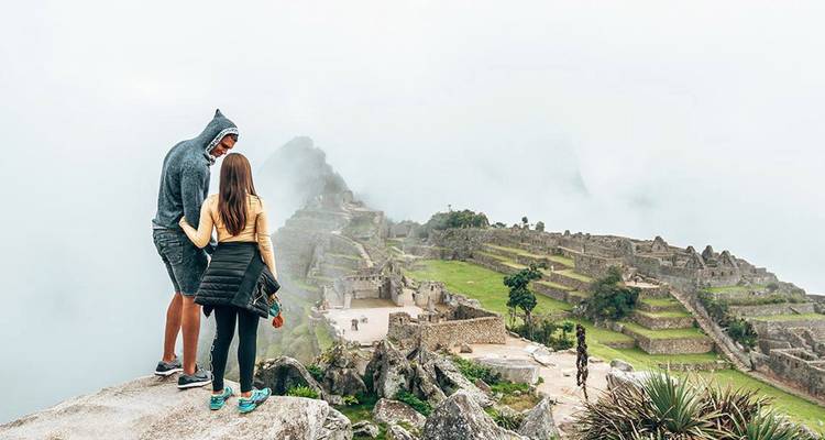 Un couple se tient sur un rebord rocheux surplombant les terrasses et ruines de Machu Picchu enveloppées de brume.