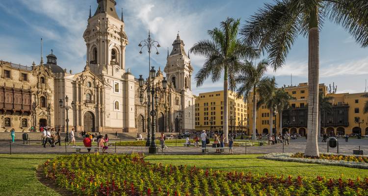 Place Mayor animée à Lima avec la Cathédrale, des bâtiments coloniaux, des palmiers et des parterres de fleurs paysagés sous un ciel bleu.