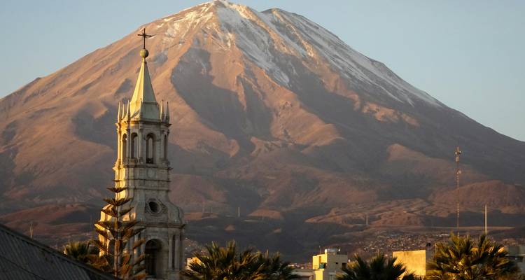A snow-capped volcano rises behind a historic church bell-tower overlooking a city at dawn light.