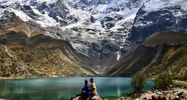Two hikers sit on a rock looking over a turquoise alpine lake framed by steep snowy mountains.