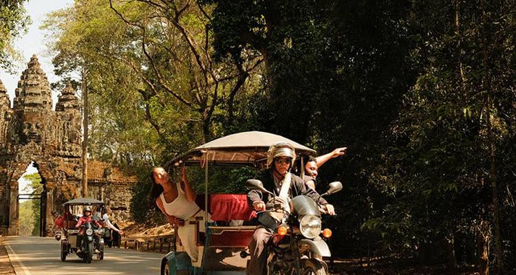 Tuk-tuks que transportan turistas pasan por la imponente puerta de piedra de Angkor Thom entre árboles de la selva.