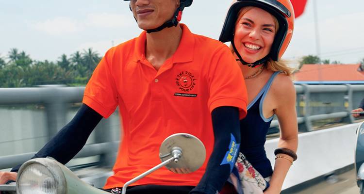 Lächelnde Frau fährt als Beifahrerin auf einem Roller, der von einem örtlichen Führer gefahren wird, über eine Brücke in Vietnam.