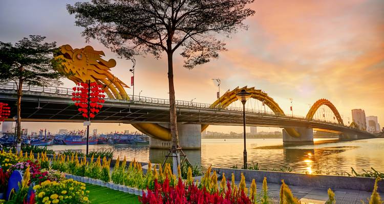 Goldene Drachenbrücke, die bei Sonnenaufgang den Fluss überspannt, mit buntem Garten im Vordergrund.