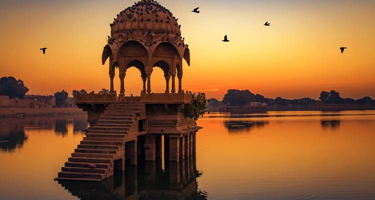 Pavillon de pierre s'élevant d'un lac tranquille au coucher de soleil orange vif avec des oiseaux silhouettés dans le ciel.