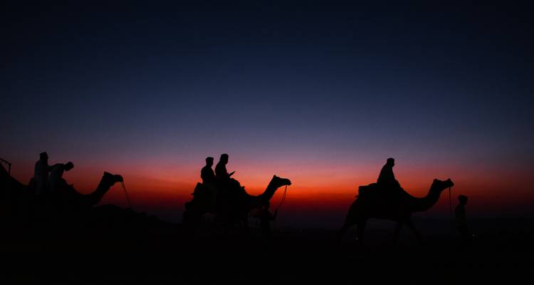 Silhouettes de chameaux et de cavaliers traversant le désert sous un ciel crépusculaire dramatique rouge et bleu.