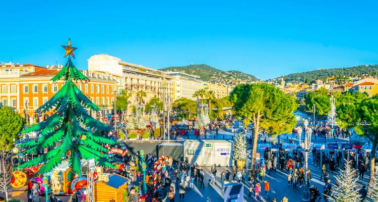 Busy Christmas market in Nice with giant decorated tree, festive stalls and crowds under bright sky.