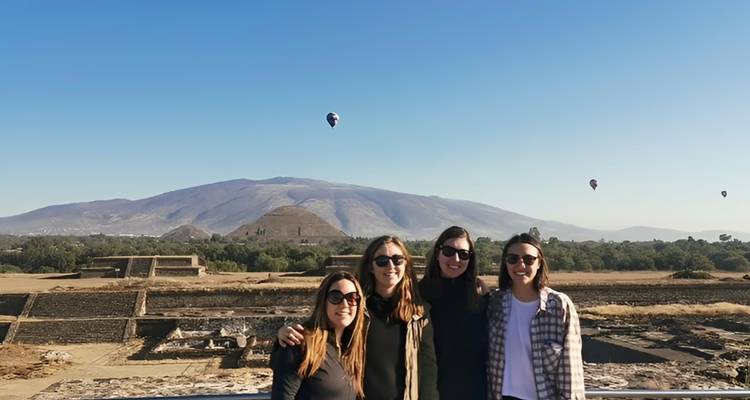 Quatre amis se tiennent bras dessus bras dessous avec la Pyramide du Soleil et des montgolfières à la dérive derrière eux.