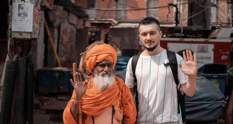 Reisender posiert mit einem orangegewandeten Sadhu auf einer Straße in Indien, beide lächeln und winken.