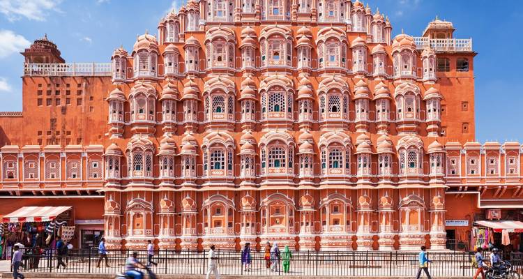 Frontale Aufnahme von Jaipurs Hawa Mahal mit Fußgängern und einem vorbeifahrenden Motorrad im Vordergrund an einem hellen Tag