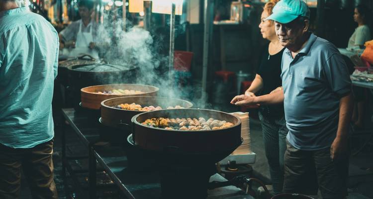 Puesto de mercado nocturno de comida donde los vendedores cuecen al vapor cestas de dumplings entre humo que se eleva