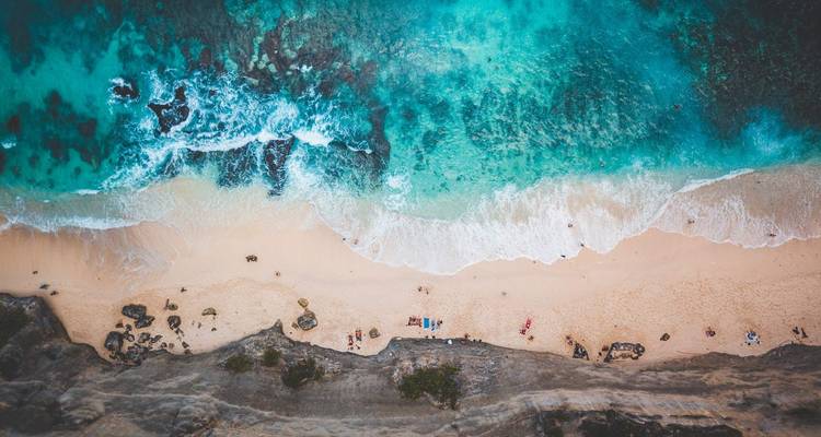 Vista aérea de dron de un océano turquesa que se encuentra con una playa de arena cremosa con olas suaves y pequeños bañistas abajo.