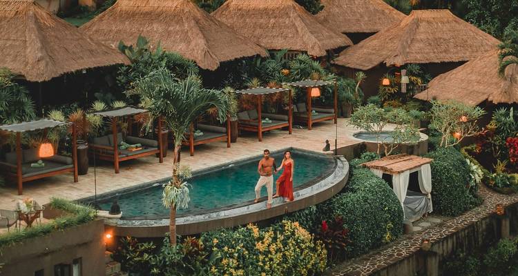 Resort selvático con techo de paja y una piscina infinita curva donde una pareja en traje de baño disfruta del exuberante entorno tropical.