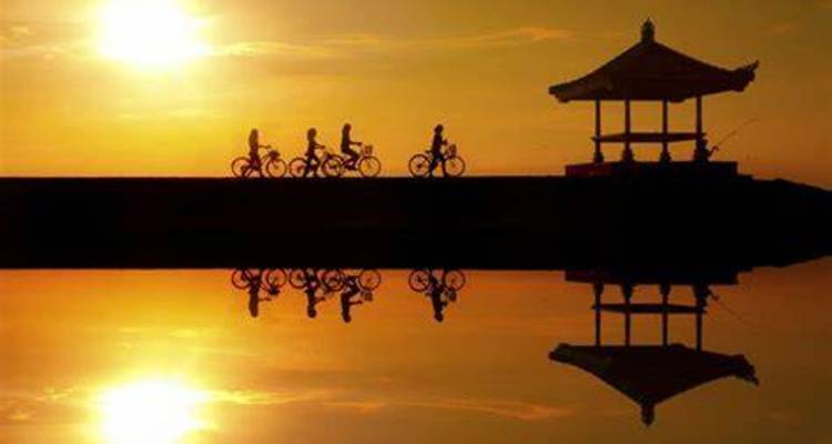 Silueta dorada del atardecer de ciclistas pasando junto a un pabellón junto al agua con reflejos perfectos en el agua quieta.