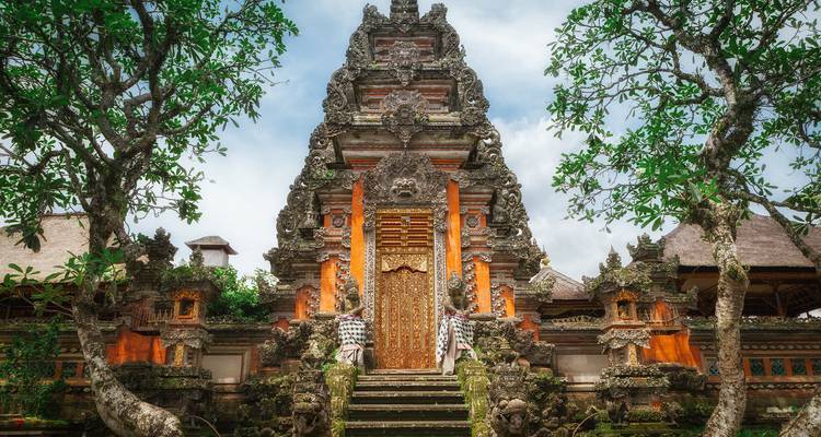 Puerta ornamentada de templo balinés con intrincados tallados en piedra enmarcada por árboles frondosos bajo un cielo brillante.