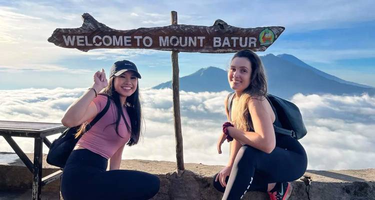 Dos excursionistas femeninas posan junto a un letrero rústico de madera que dice 'Bienvenidos al Monte Batur' sobre un mar de nubes.