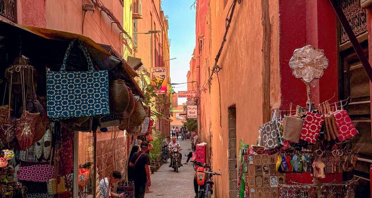 Narrow ochre alley in Marrakesh souk lined with colorful textiles, shops and locals under warm light.
