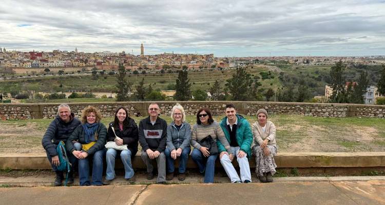 Tour group seated on a low wall with a panoramic view of an historic Moroccan city under an overcast sky.