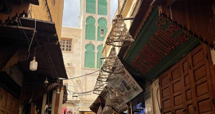 Covered lane in Fes medina with traditional wooden balconies and a tall green-tiled minaret rising above.