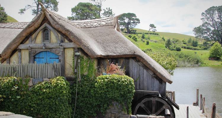 Charming thatched millhouse with blue door and water wheel beside a tranquil lake at the Hobbiton movie set.