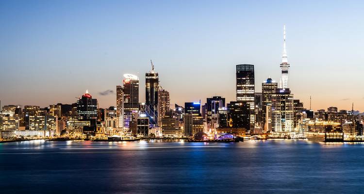 Evening view of illuminated Auckland CBD skyscrapers reflecting across calm harbour waters.