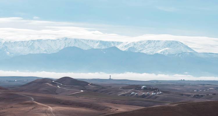 Extensa llanura desértica con senderos serpenteantes y las nevadas montañas del Atlas alzándose sobre la capa de nubes.