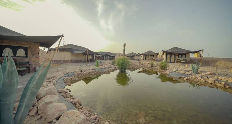 Campamento de glamping en el desierto con tiendas alrededor de un pequeño estanque reflectante bajo un cielo brumoso.