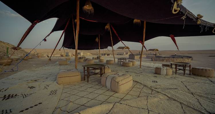 Tienda de descanso nómada sombreada con mesas bajas y pufs sobre alfombras estampadas en el desierto.