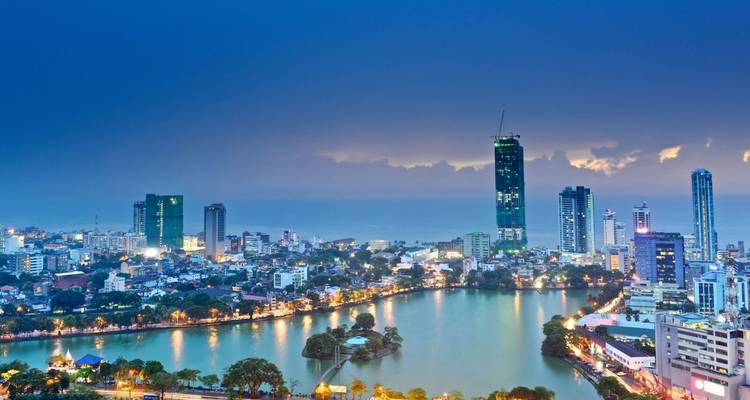 Panorama du soir de l'horizon de Colombo avec des tours de grande hauteur et les lumières de la ville se reflétant sur le lac Beira sous un ciel bleu profond.