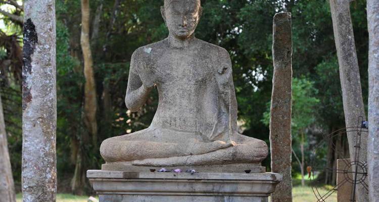 Bouddha de pierre patiné par les intempéries assis en méditation à l'extérieur, encadré par des ruines boisées.