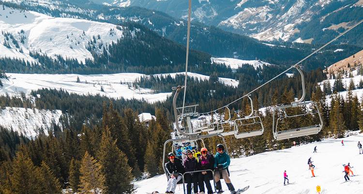Skiers ride an alpine chairlift above snowy pistes and conifer forests in the Austrian Tyrol mountains.