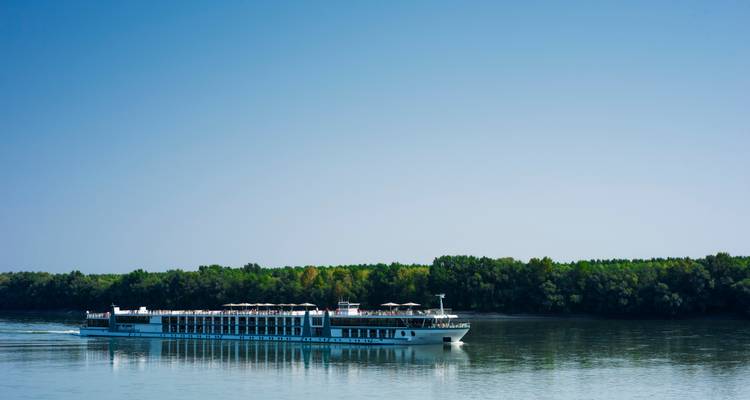 River cruise ship sailing calmly past a forested shoreline under a clear blue sky