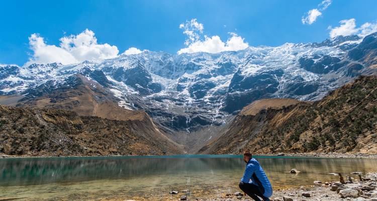 Lago turquesa cristalino bajo imponentes picos cargados de nieve mientras un caminante solitario se arrodilla en la orilla rocosa.