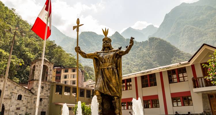 Statue en bronze d'un roi Inca sur une place avec le drapeau péruvien et des montagnes vertes brumeuses en arrière-plan.