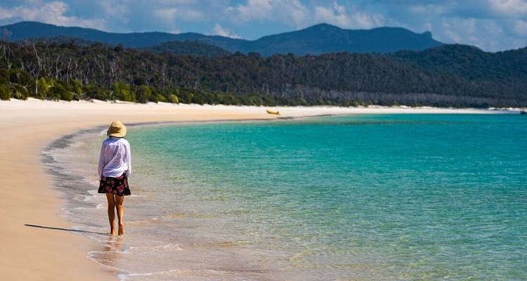 Femme au chapeau de soleil se promène le long d'une plage de sable blanc immaculé bordée d'eaux turquoise et de promontoires boisés.