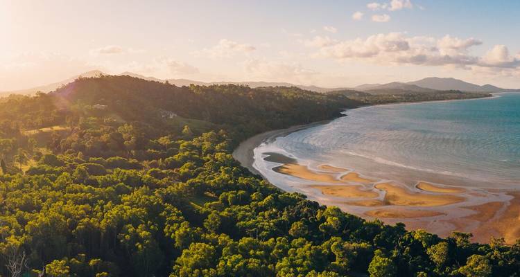 Vue aérienne à l'heure dorée d'un littoral luxuriant, d'une plage sinueuse et de collines boisées rencontrant une mer calme.
