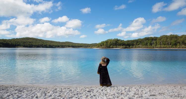 Un voyageur se tient sur le sable blanc en regardant les eaux bleues cristallines d'un lac d'eau douce entouré de forêt.