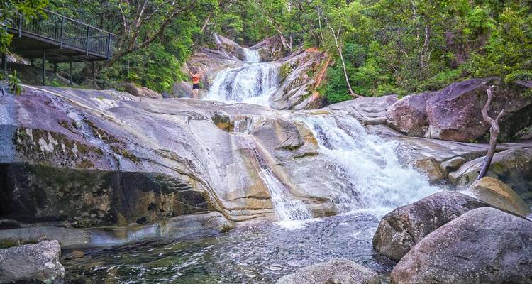 Cascade à plusieurs niveaux qui dévale sur des dalles de granit dans une forêt tropicale luxuriante ; un randonneur observe depuis une passerelle.