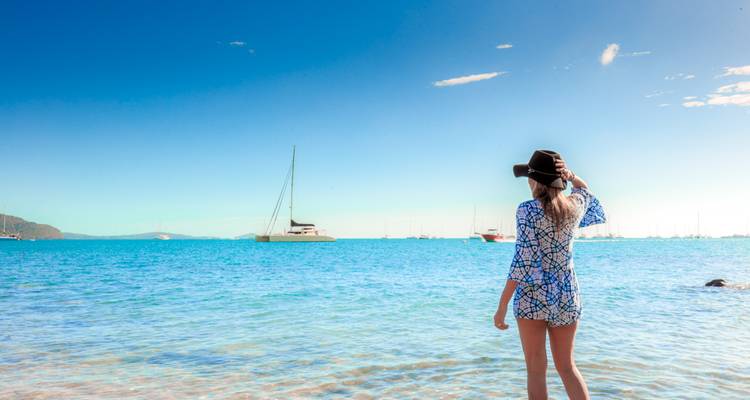 Vrouw in gedessineerde jumpsuit staat kniediep in turkooizen baai kijkend naar geankerde jachten onder heldere blauwe hemel.