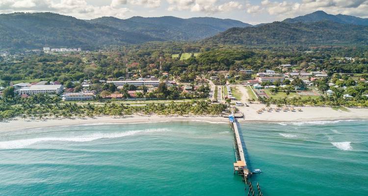 Drone view of a coastal Honduran town, sandy beach, pier and lush mountains rising behind.